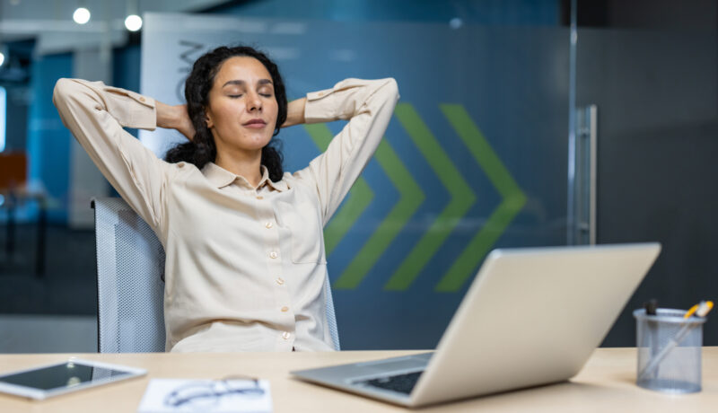 Hispanic businesswoman relaxing at desk with eyes closed in modern office setting